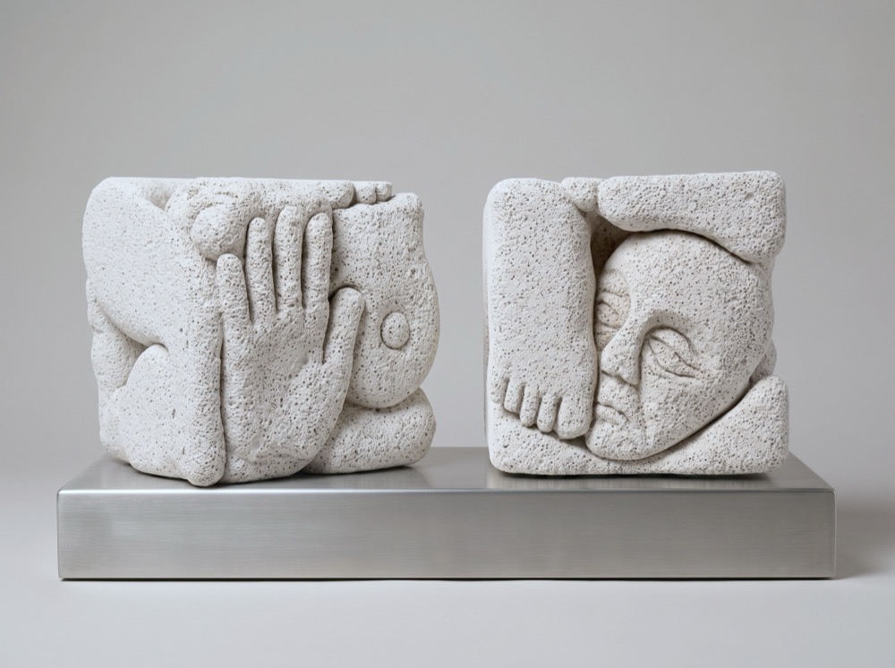Two small aerated-concrete cubes side by side on a long steel plinth. An open hand presses against the face of the left cube; a face in profile and a foot are pressed into the right one.