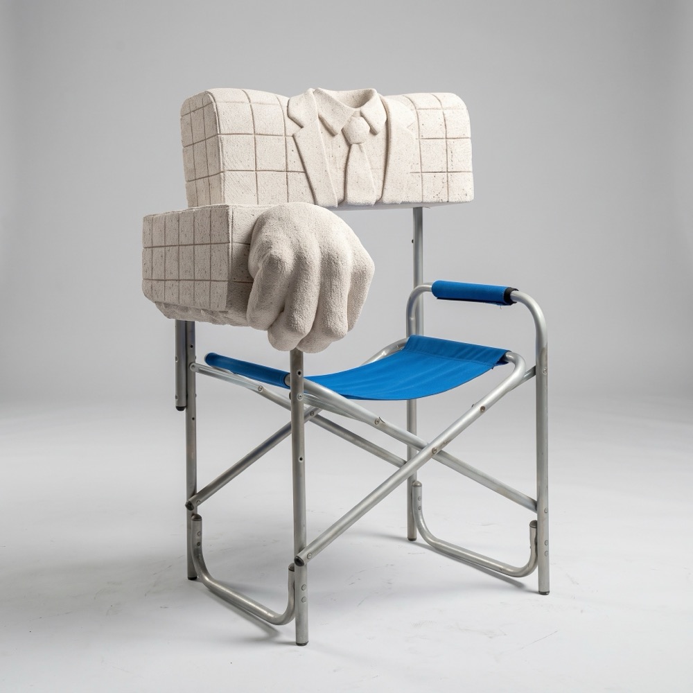 A folding director’s chair of aluminium and blue fabric. Its backrest is an aerated-concrete torso in a checked suit and tie, and a concrete hand grips the armrest.