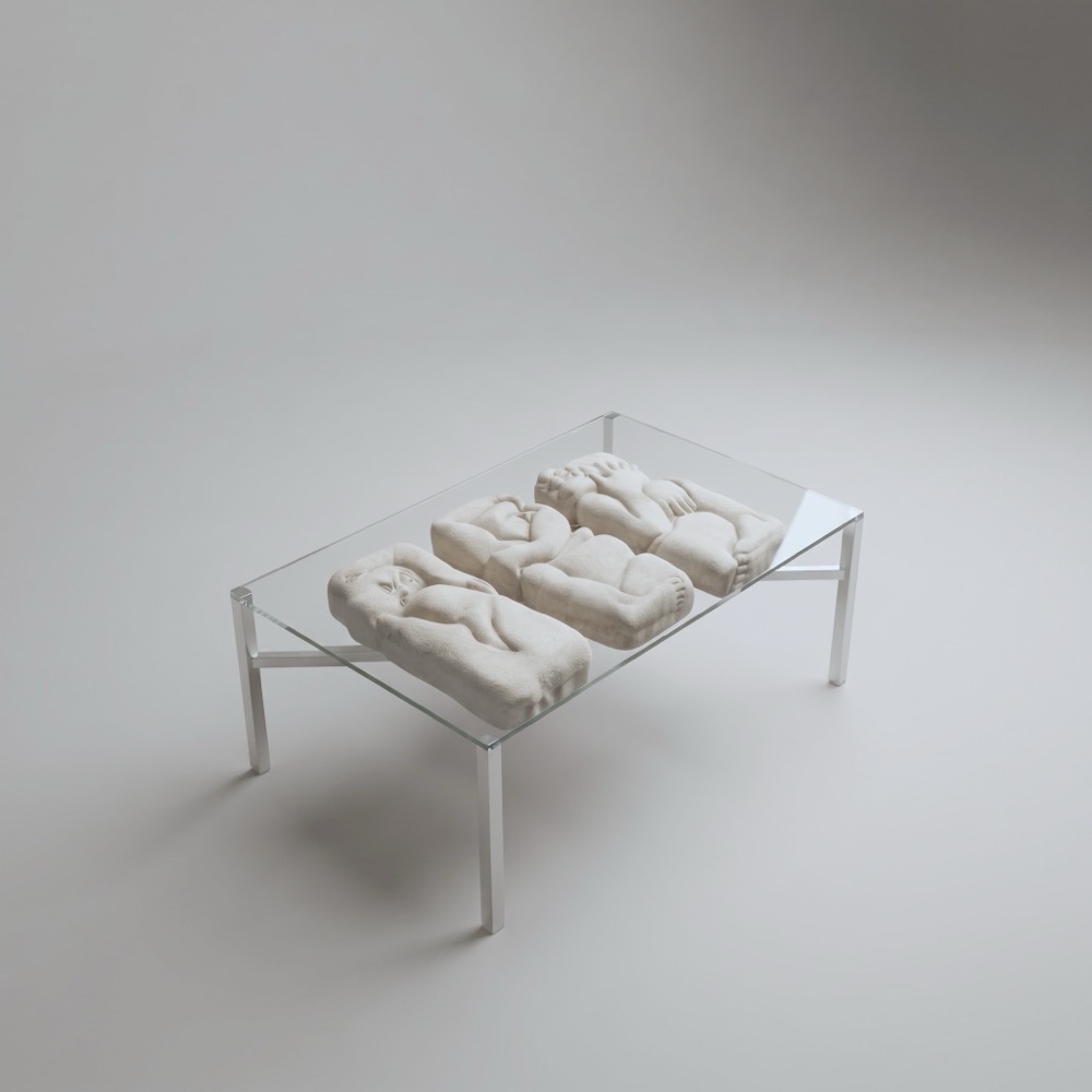 A low table with a glass top on thin steel legs. Three aerated-concrete slabs lie in a row on the glass, each carrying a reclining figure in relief.